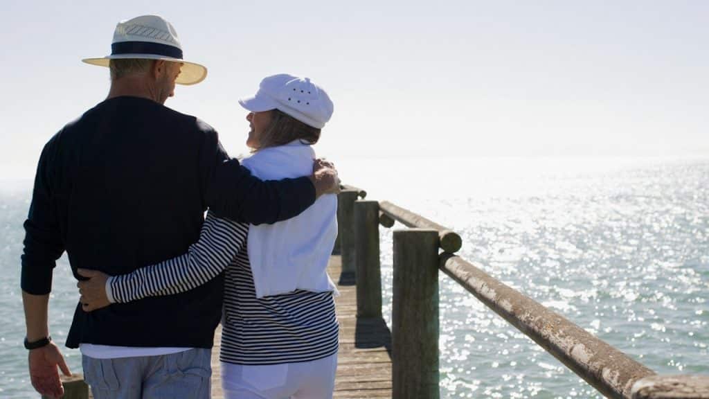 A couple walking down a pier with their arms around each other’s backs and smiling at each other.