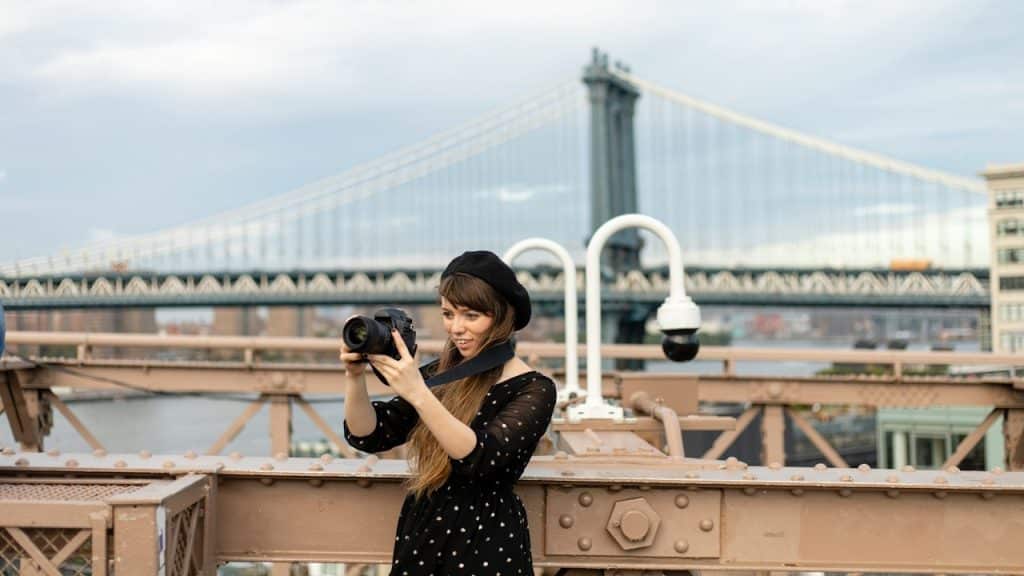 A woman holding a camera is taking a picture in front of a bridge.