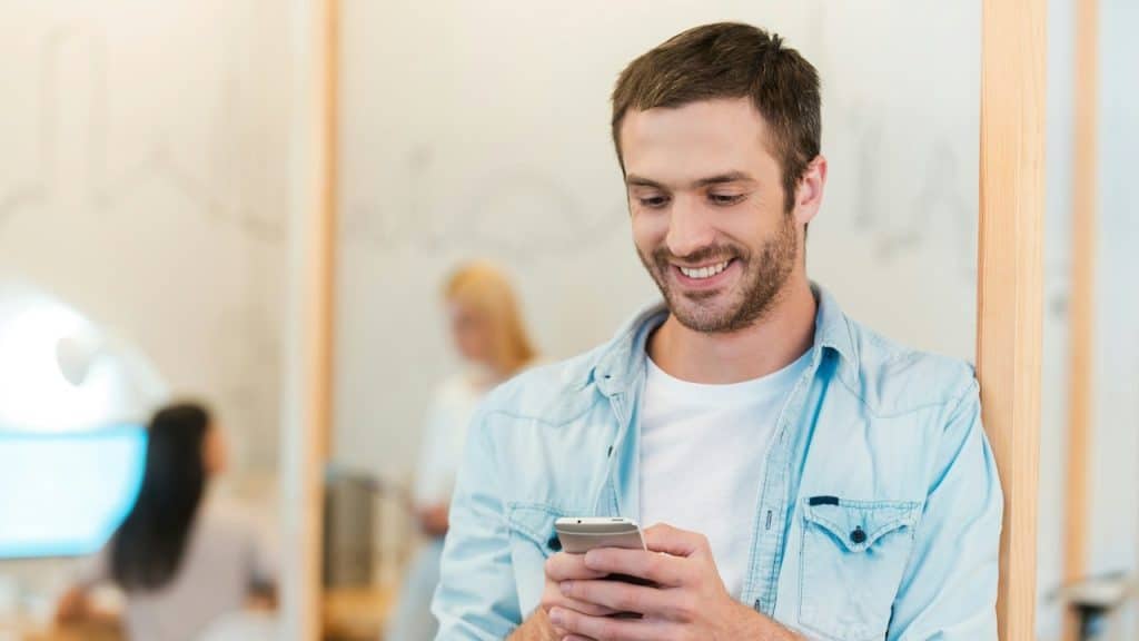 Happy young man looking at his mobile phone and smiling while his colleagues are working in the background.
