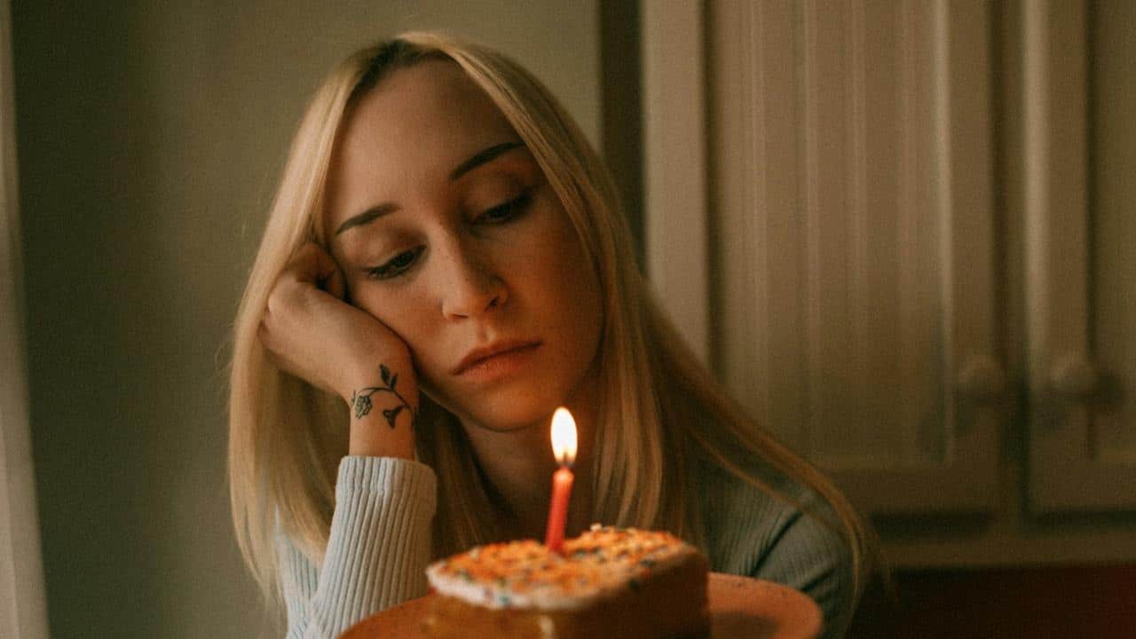 A woman holding a cake with a candle on it sitting alone, appearing sad.