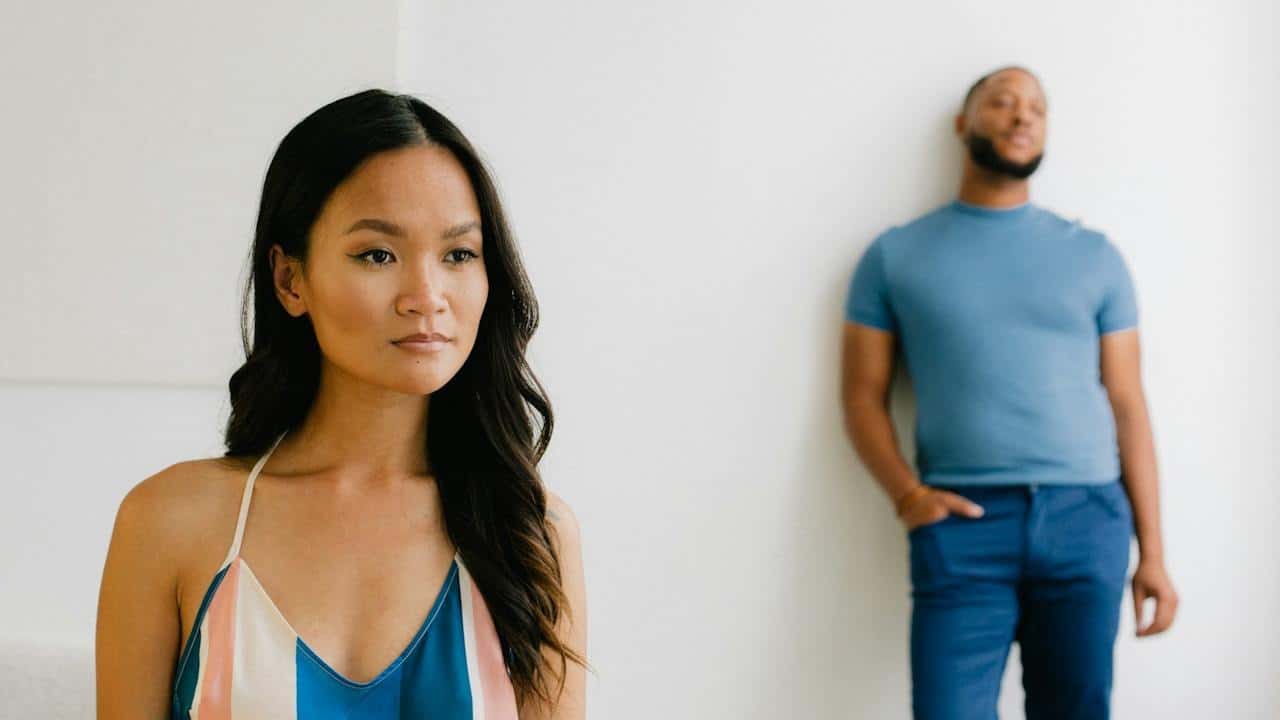A woman is standing in the forefront while a bearded man leans against a wall in the background.