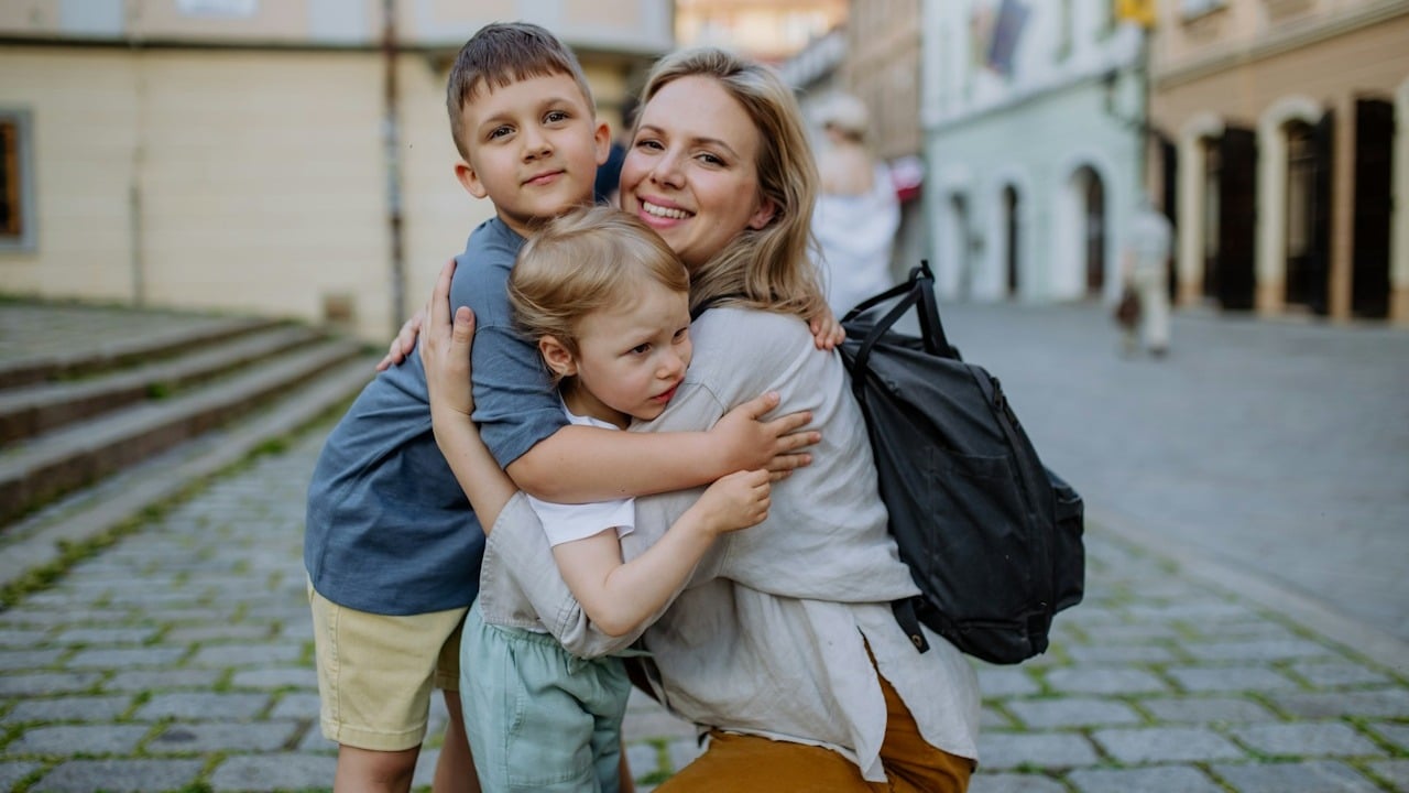 A young mother and little children smiling confidently and hugging each other while standing at a street.