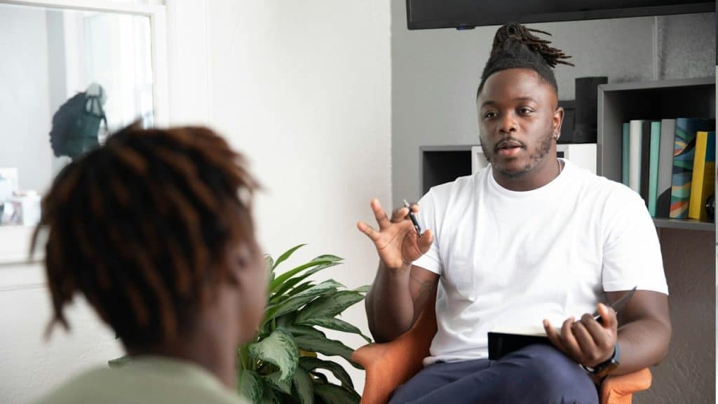 A lightly bearded man with dreadlocks is talking to a woman while sitting on a chair.