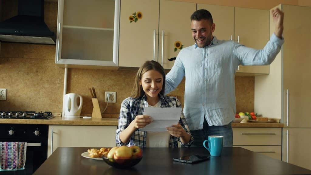 A woman is reading a document while a man is extending his arms with joy.