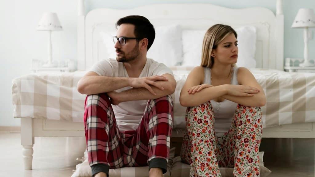 A couple sitting on the floor and leaning against the bed while looking in opposite directions.