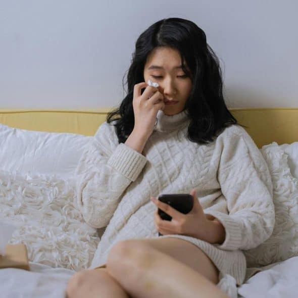 A woman sitting on a bed, wiping her tears while looking at her phone.