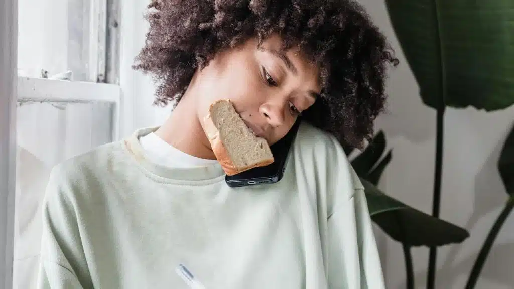 A woman holding a phone with her shoulder while a piece of bread rests against her cheek.