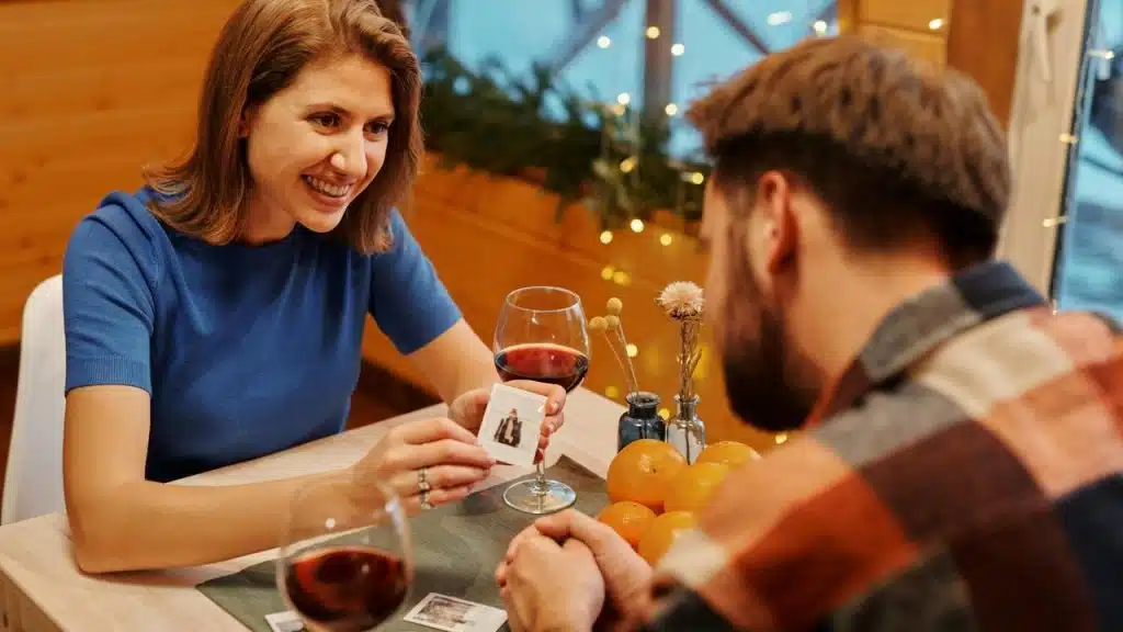 A woman smiling and showing a small photo to a man across a table with glasses of wine.