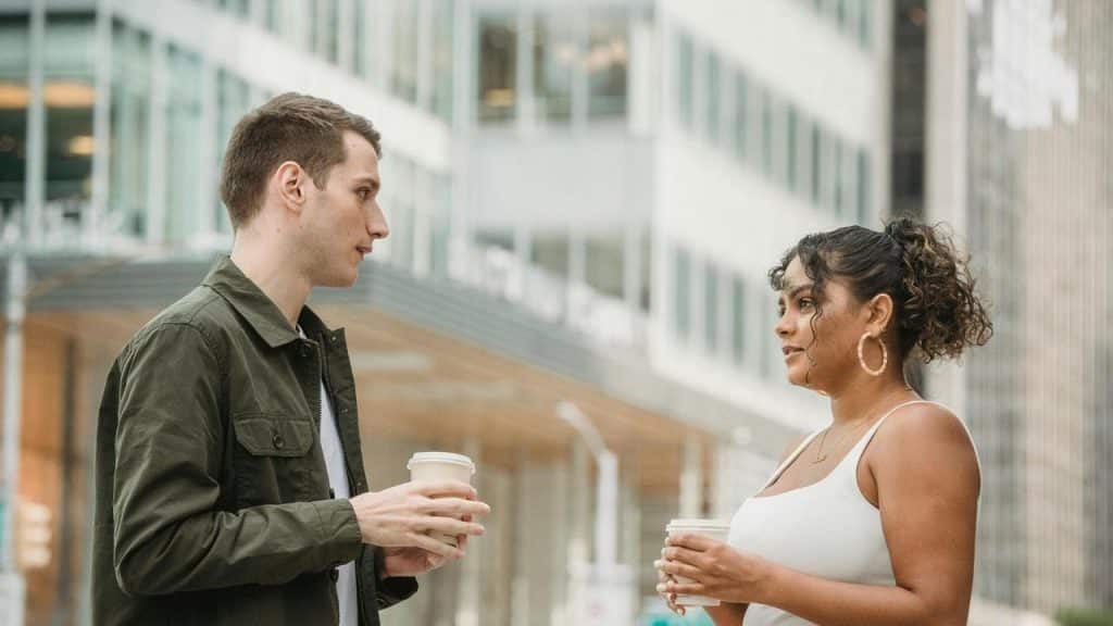 A man and woman standing outside with coffee cups, talking in front of an office building.