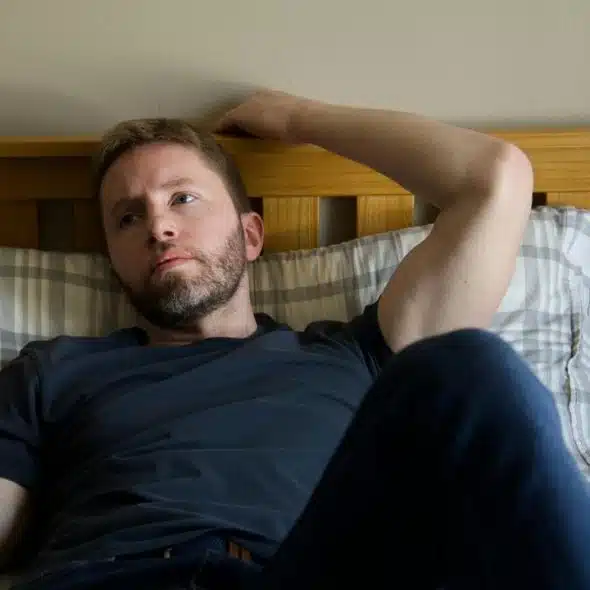 A man lying on a bed with his hand behind his head, gazing thoughtfully to the side.