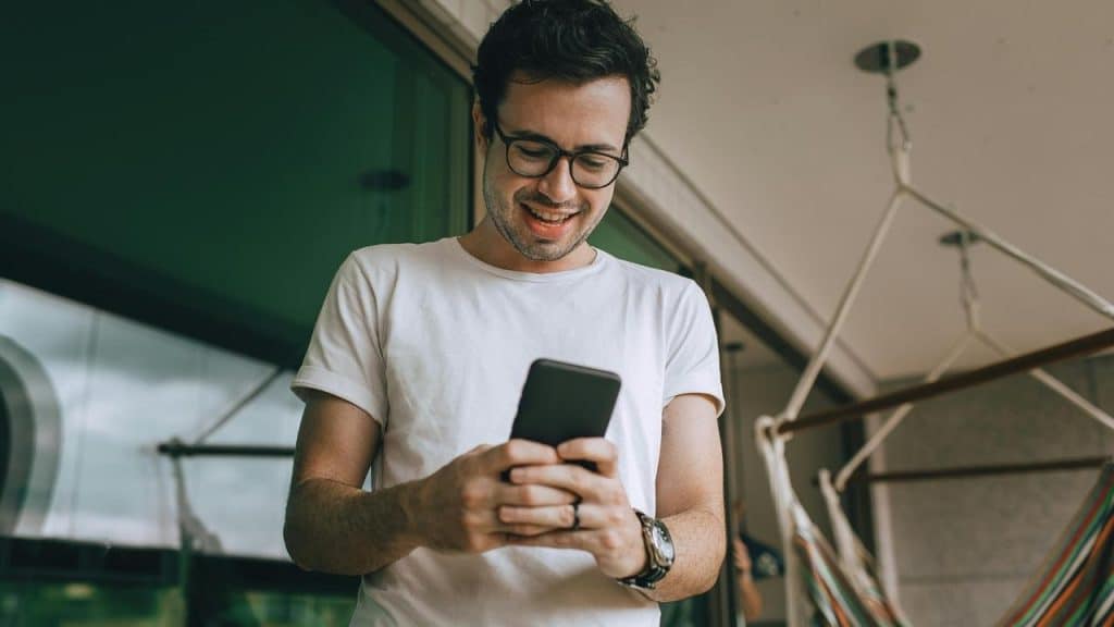 A smiling man wearing glasses looking at his phone indoors.