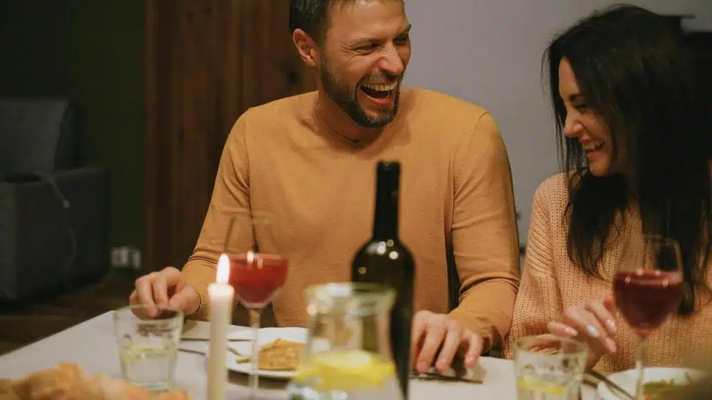 A couple laughing together at a candlelit dinner with wine and food on the table.