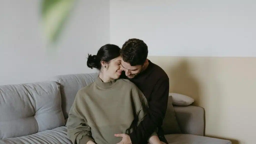 A couple sitting on a couch, gently embracing and smiling with their heads touching.