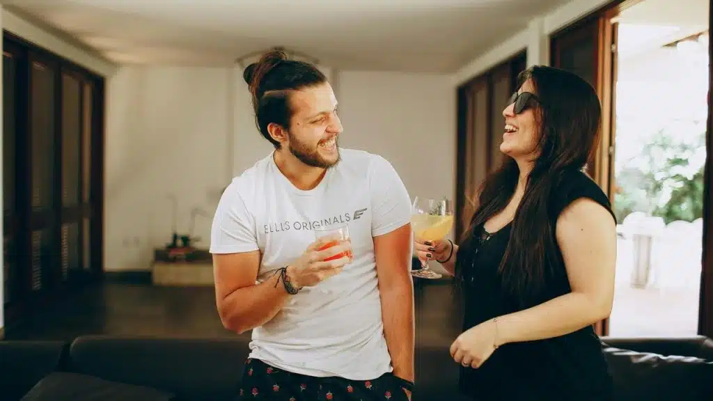 A man and woman laughing while holding drinks in a bright living room.