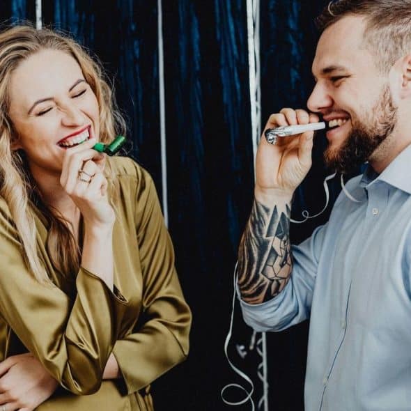 A smiling couple laughing and blowing party horns in front of a dark festive backdrop.
