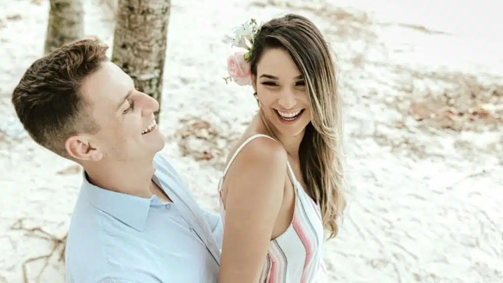 A smiling couple embracing on a sandy beach, with a flower in the woman’s hair.