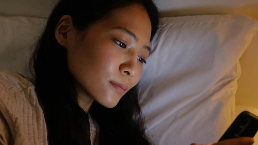 A woman lying in bed looking at her phone in warm light.