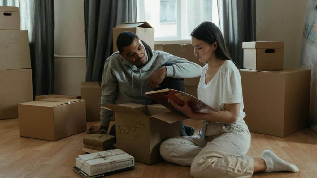 A couple sitting on the floor surrounded by moving boxes, looking through books.