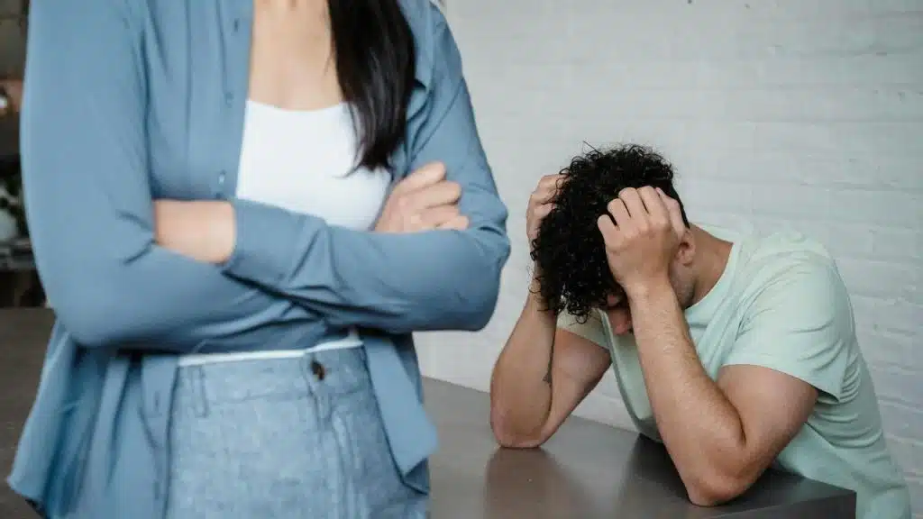 A man holding his head while a woman stands with her arms crossed.