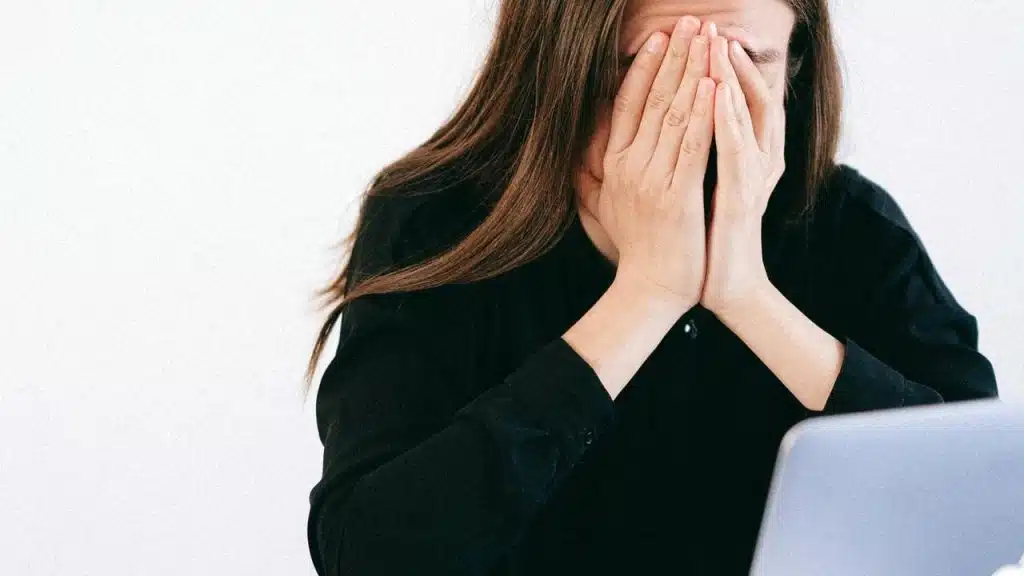 A woman covering her face while sitting at a desk with a laptop and papers.