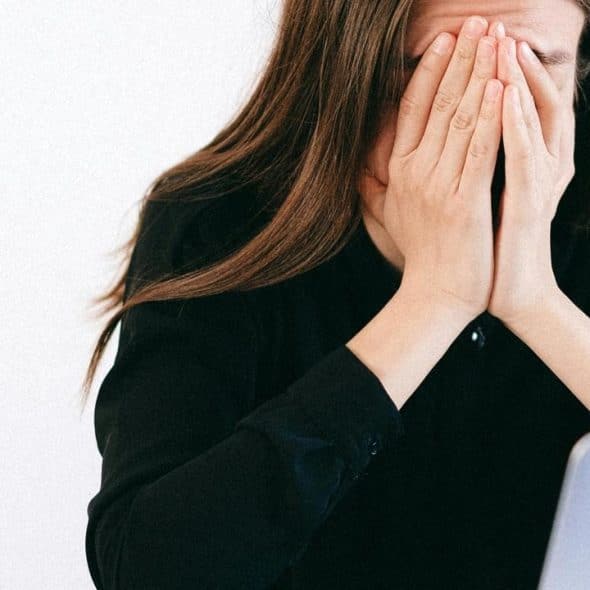 A woman covering her face while sitting at a desk with a laptop and papers.
