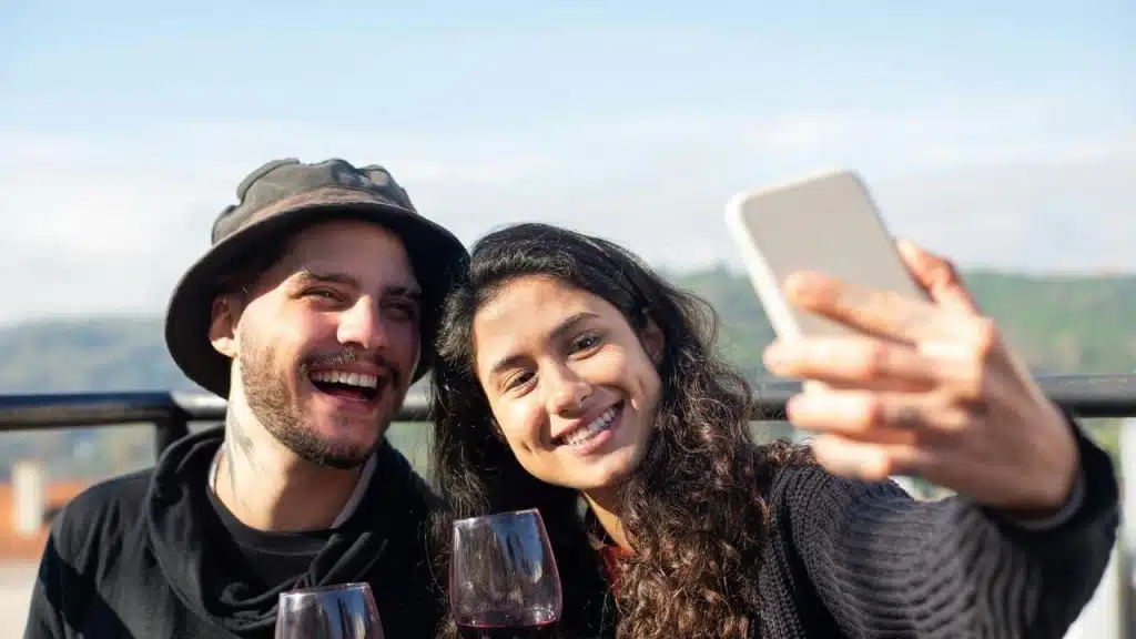 A smiling couple taking a selfie while holding glasses of wine outdoors.
