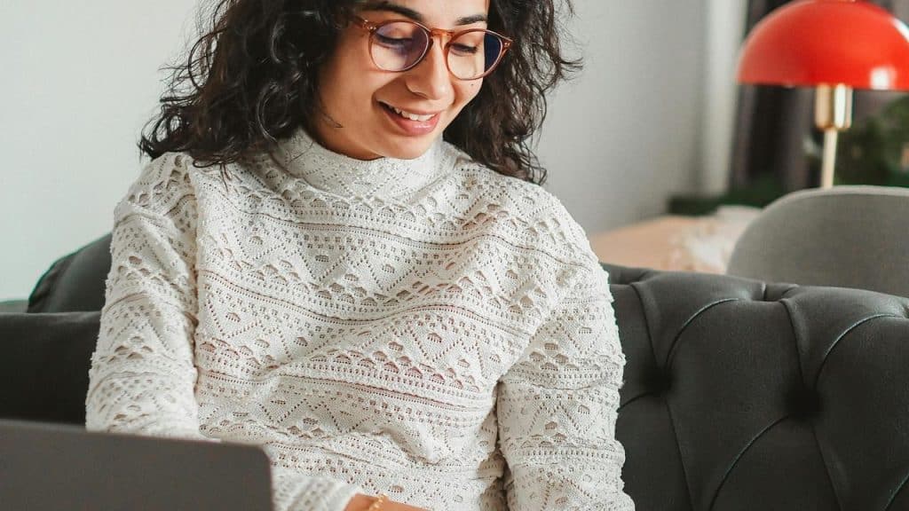A smiling woman wearing glasses while using a laptop on a couch.