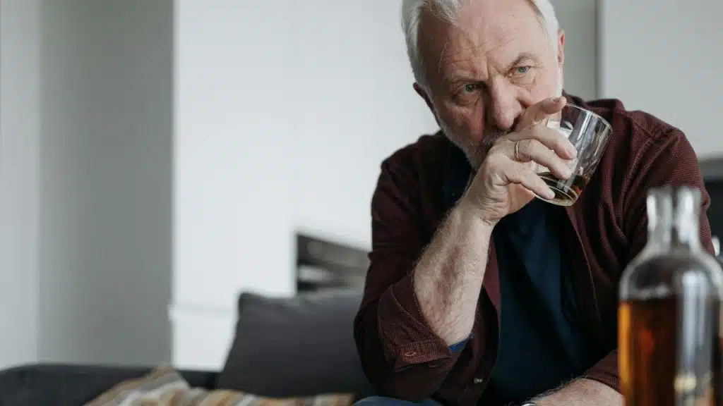 A man with gray hair sits on a couch and drinks from a glass tumbler.