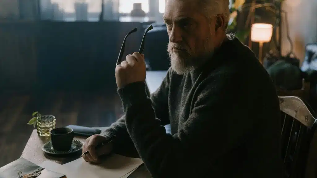 A man with a gray beard holds glasses while sitting at a desk with paper.