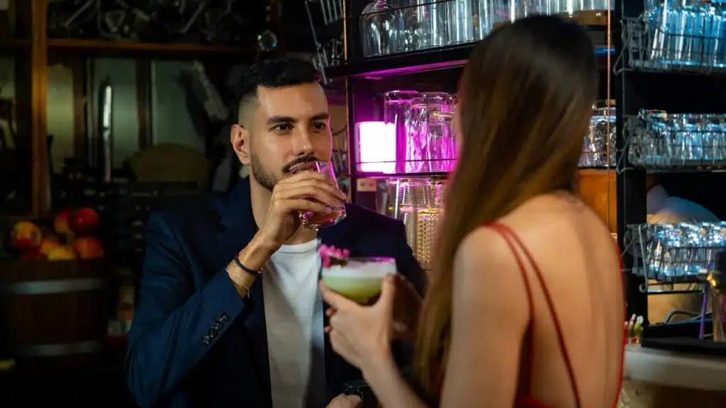A man in a dark blazer drinks from a glass while facing a woman indoors.