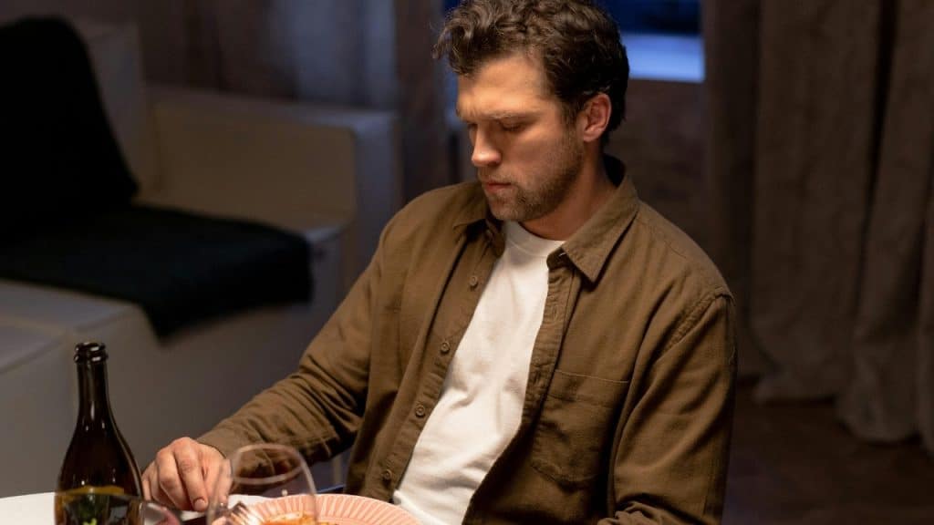 A man in a brown shirt sits at a dining table looking down at a plate.