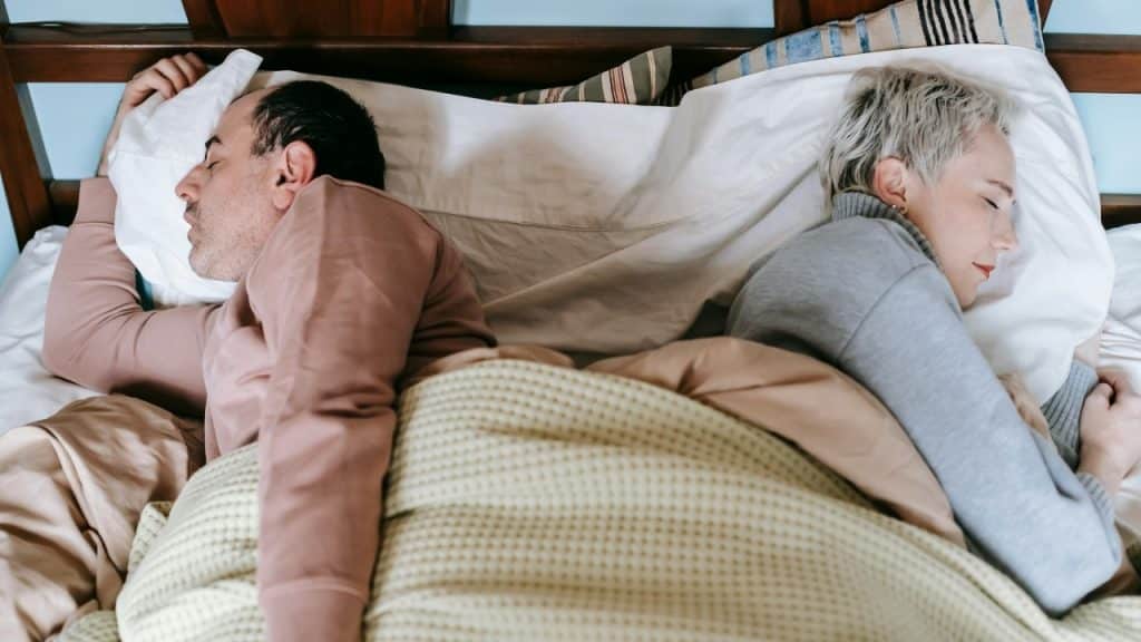 Two people lie in bed with their backs to each other under a thick blanket.