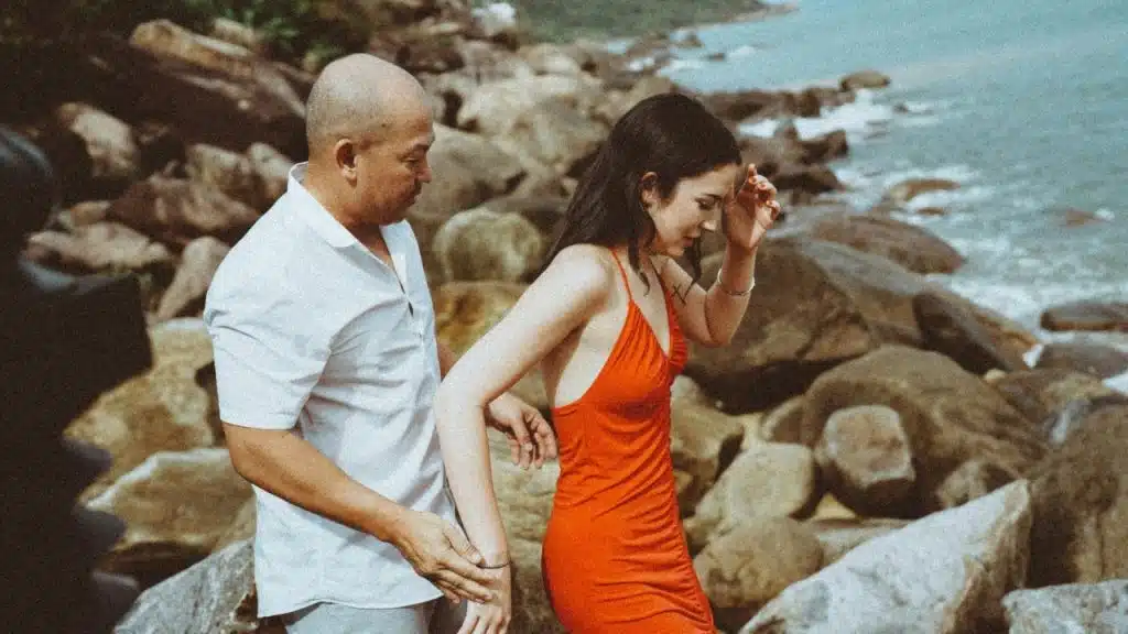 A man in a white shirt assists a woman in a red dress over coastal rocks.