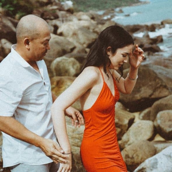 A man in a white shirt assists a woman in a red dress over coastal rocks.