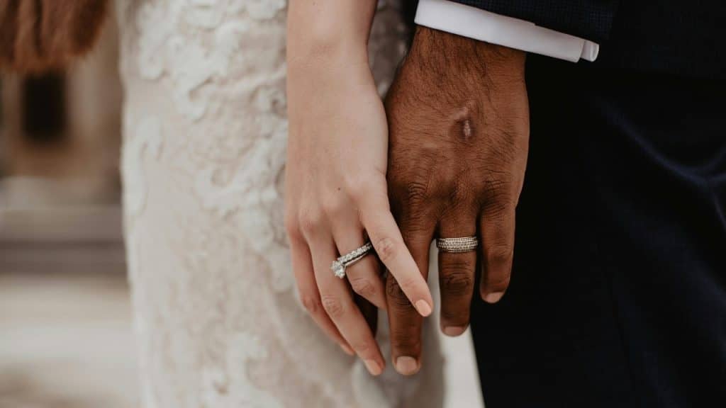 A close-up of two people holding hands, showcasing a diamond engagement ring and a wedding band.