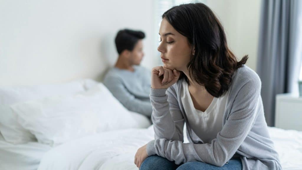 A woman sitting on the edge of a bed with a blurred figure in the background.