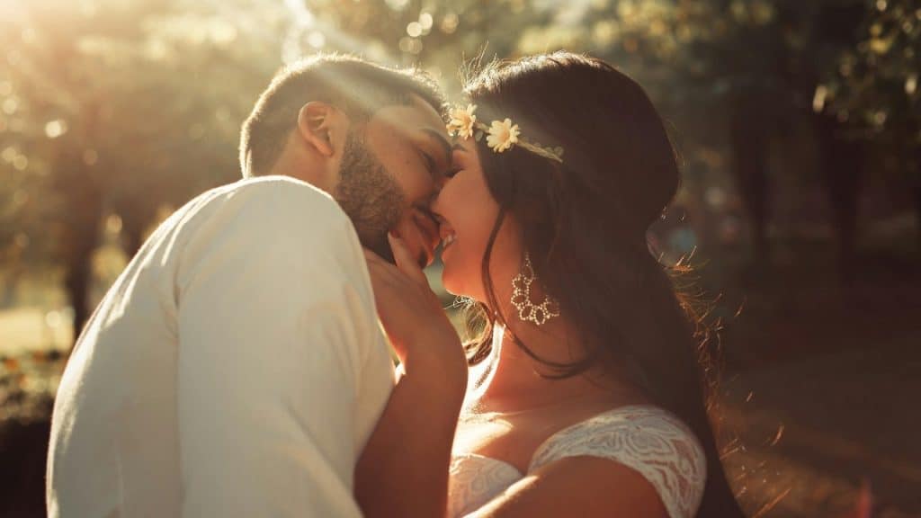 Two people with their faces close together outdoors with sunlight filtering through trees in the background.