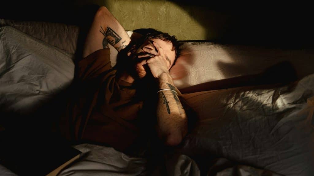 A man lying in bed with his hand covering his face in a dark, shadowed room.