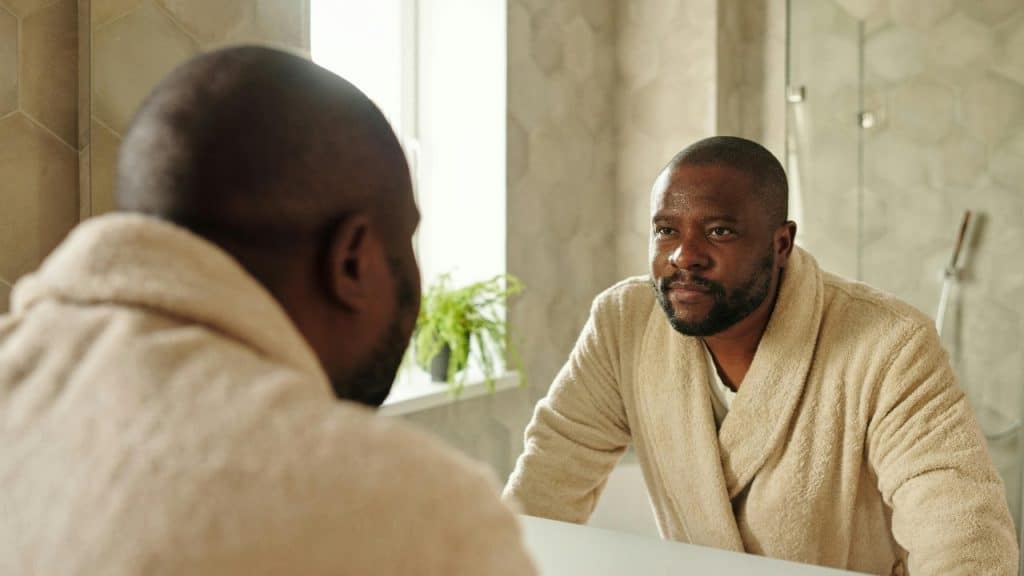 A man wearing a beige robe looking at his own reflection in a bathroom mirror.