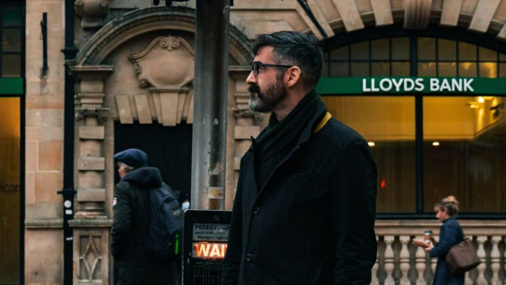 A bearded man in a dark coat standing on a city street near a bank.