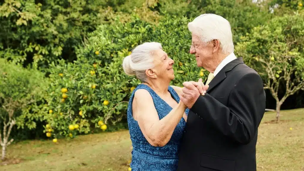 An elderly couple in formal attire smiling and dancing together in an outdoor garden setting.