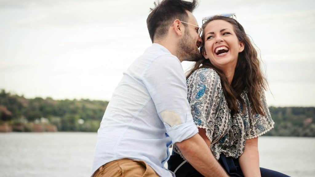 A man is about to kiss a laughing woman on the cheek while they sit in front of a lake.