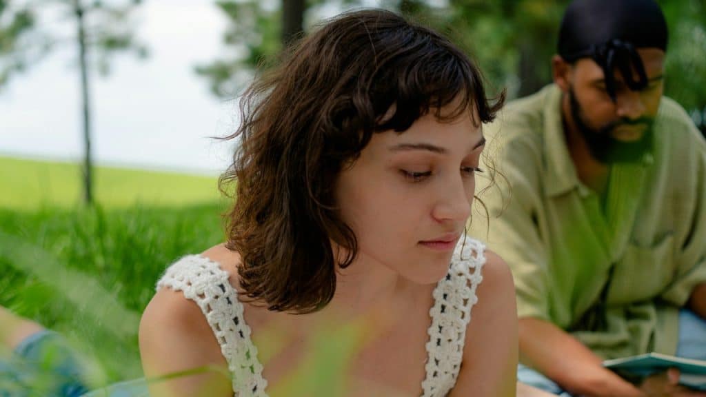 A woman looking downwards while a bearded man sits behind her.