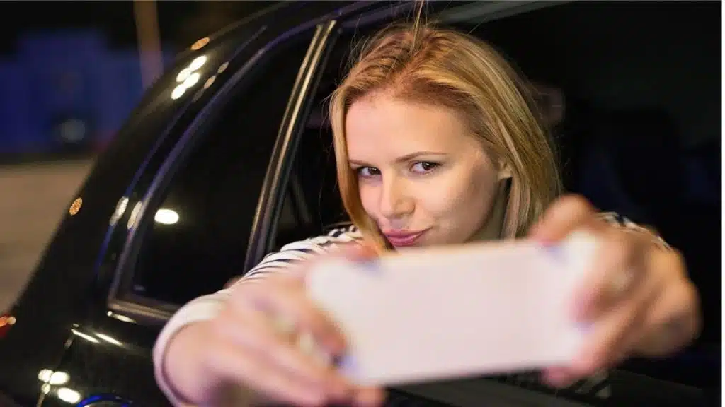 A woman in a city in her modern car at night holding a smartphone and taking a selfie.