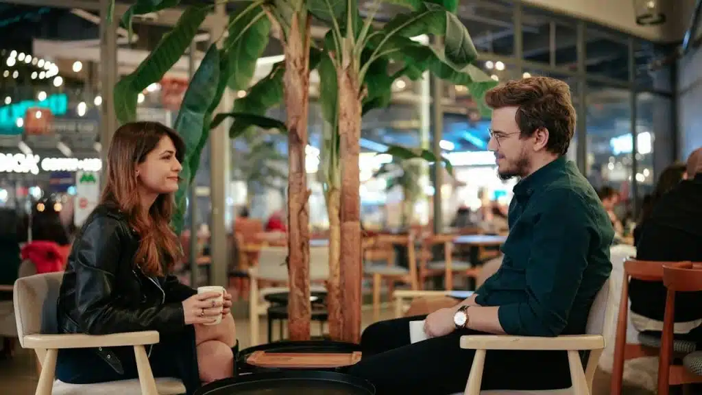 A couple is seated, facing each other in a brightly lit cafe or indoor seating area with plants, having a conversation over coffee.