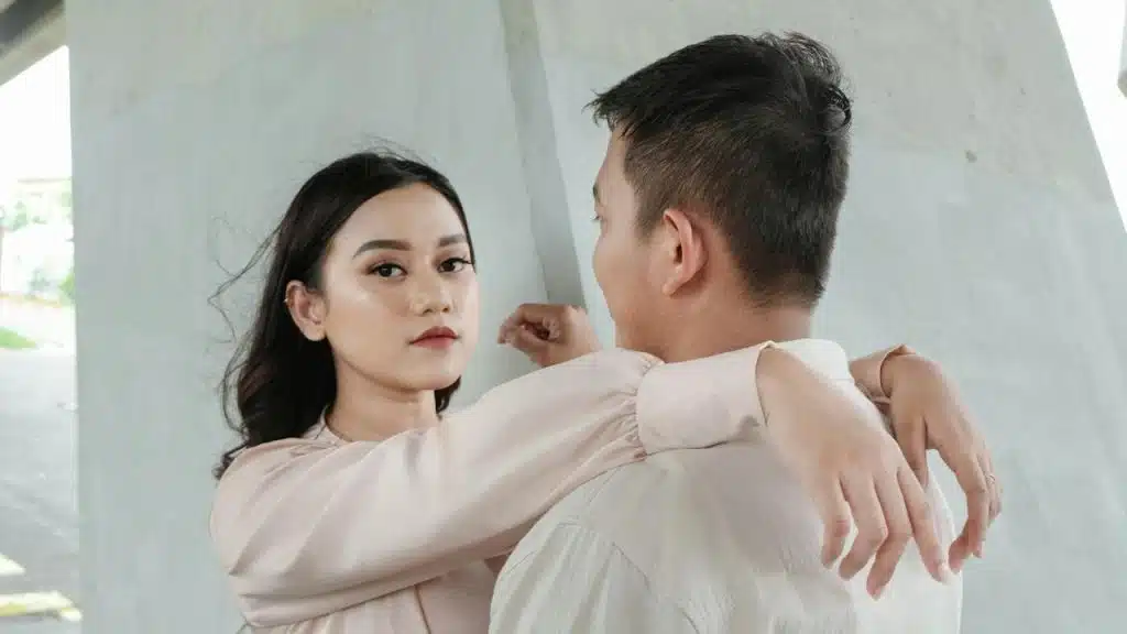 A woman with an intense expression has her arms wrapped around a man's neck as she looks directly at the camera.