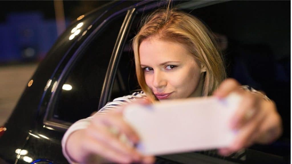 A woman in a city in her modern car at night holding a smartphone and taking a selfie.