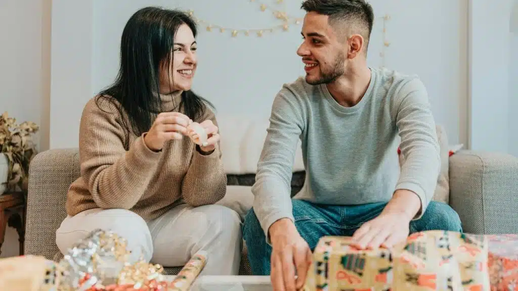 A couple smiling while wrapping gifts together on a couch.