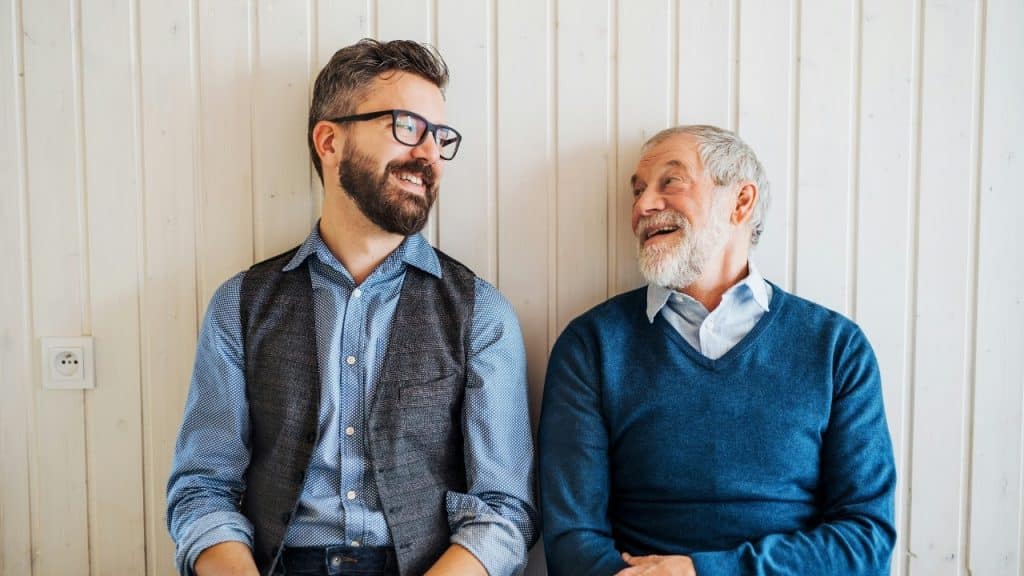 Two men with beards sitting against a white wood-paneled wall, smiling and looking at each other.