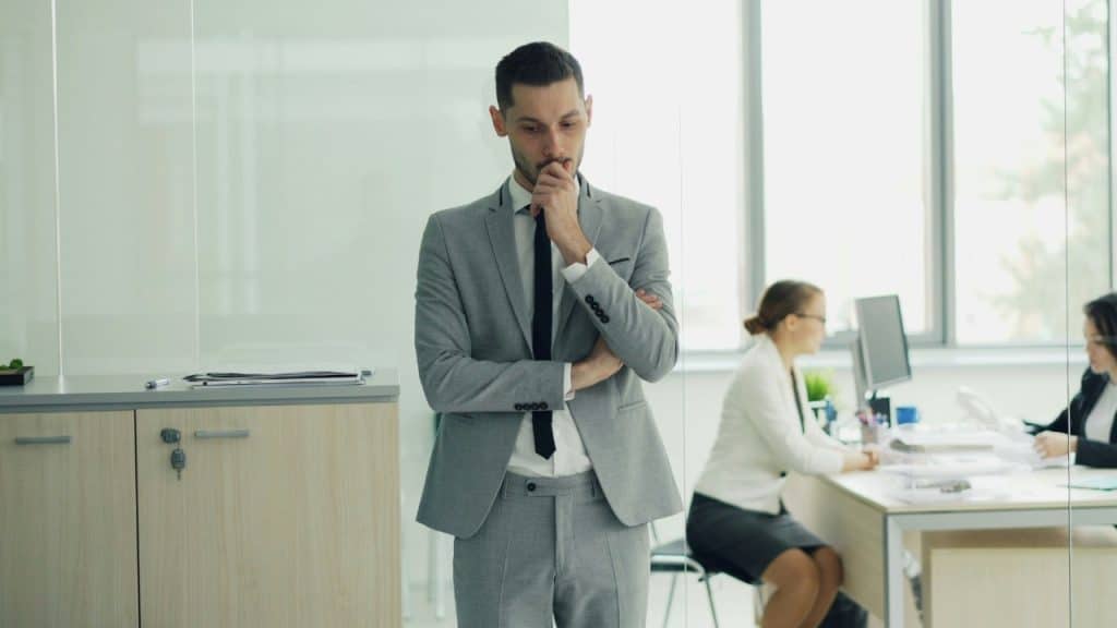 A man in a grey suit standing in an office, looking down thoughtfully with hand on chin.